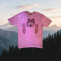 A heather pink short-sleeve t-shirt featuring a graphic print of a wolf's face centered between two dreamcatcher designs, displayed against a neutral gray gradient background.